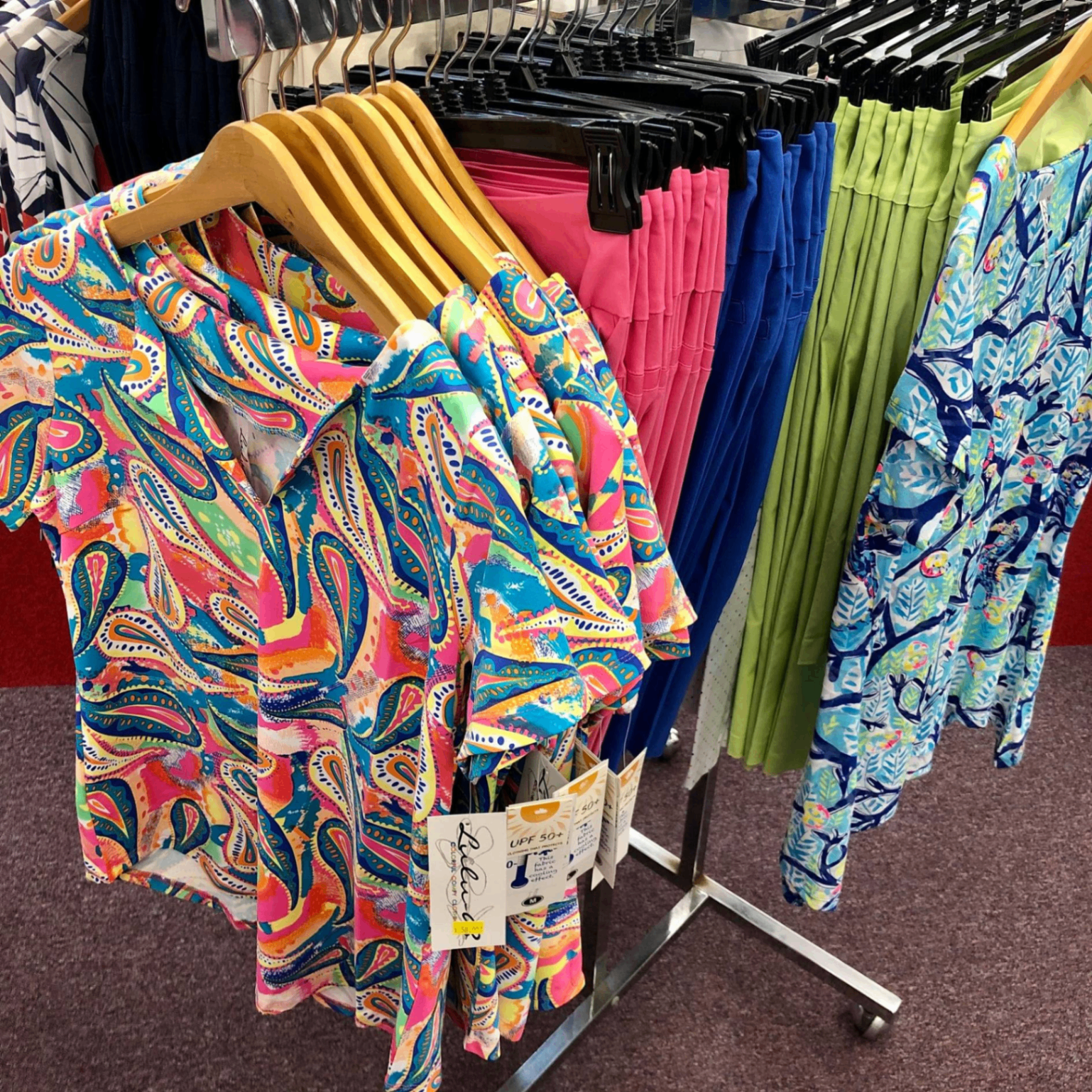 Lulu-B brightly colored paisley camp shirt with UPF 50+ tag, hanging next to pink, blue, and green pull-on skirts