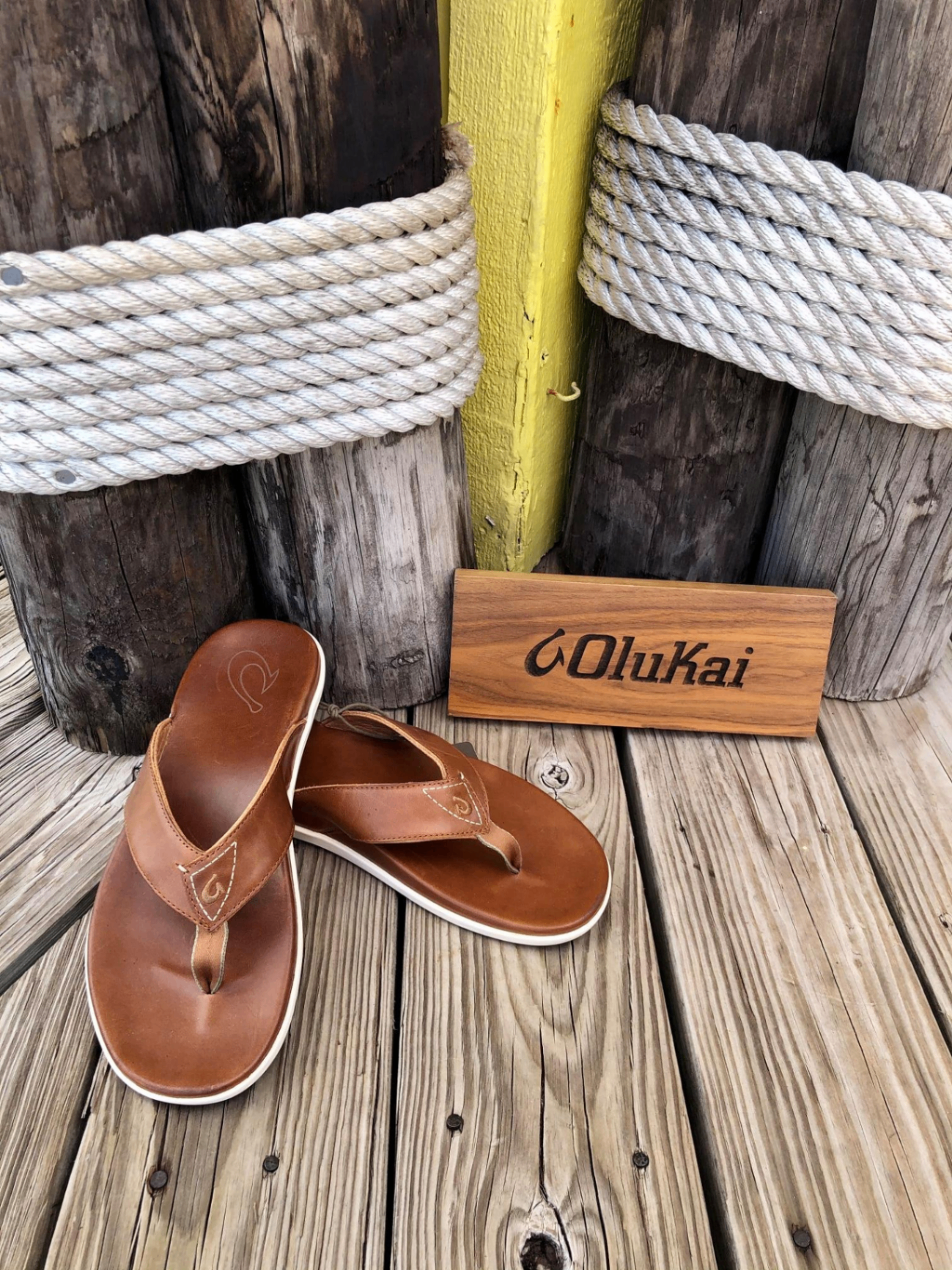 OluKai brown leather flip flops displayed on a weathered wood dock with rope detail