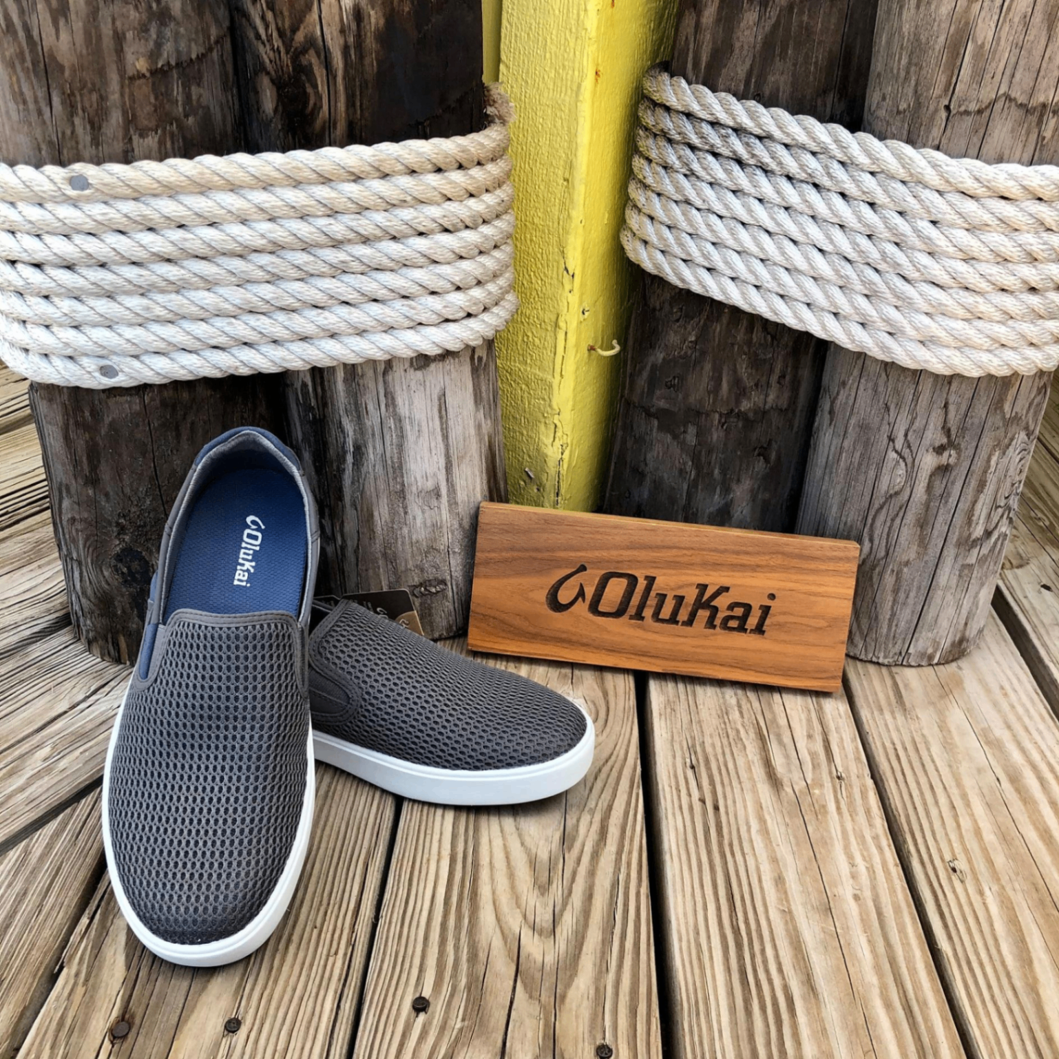 OluKai Pehuea grey mesh slip-on sneakers on a wooden dock beside a yellow piling wrapped in rope