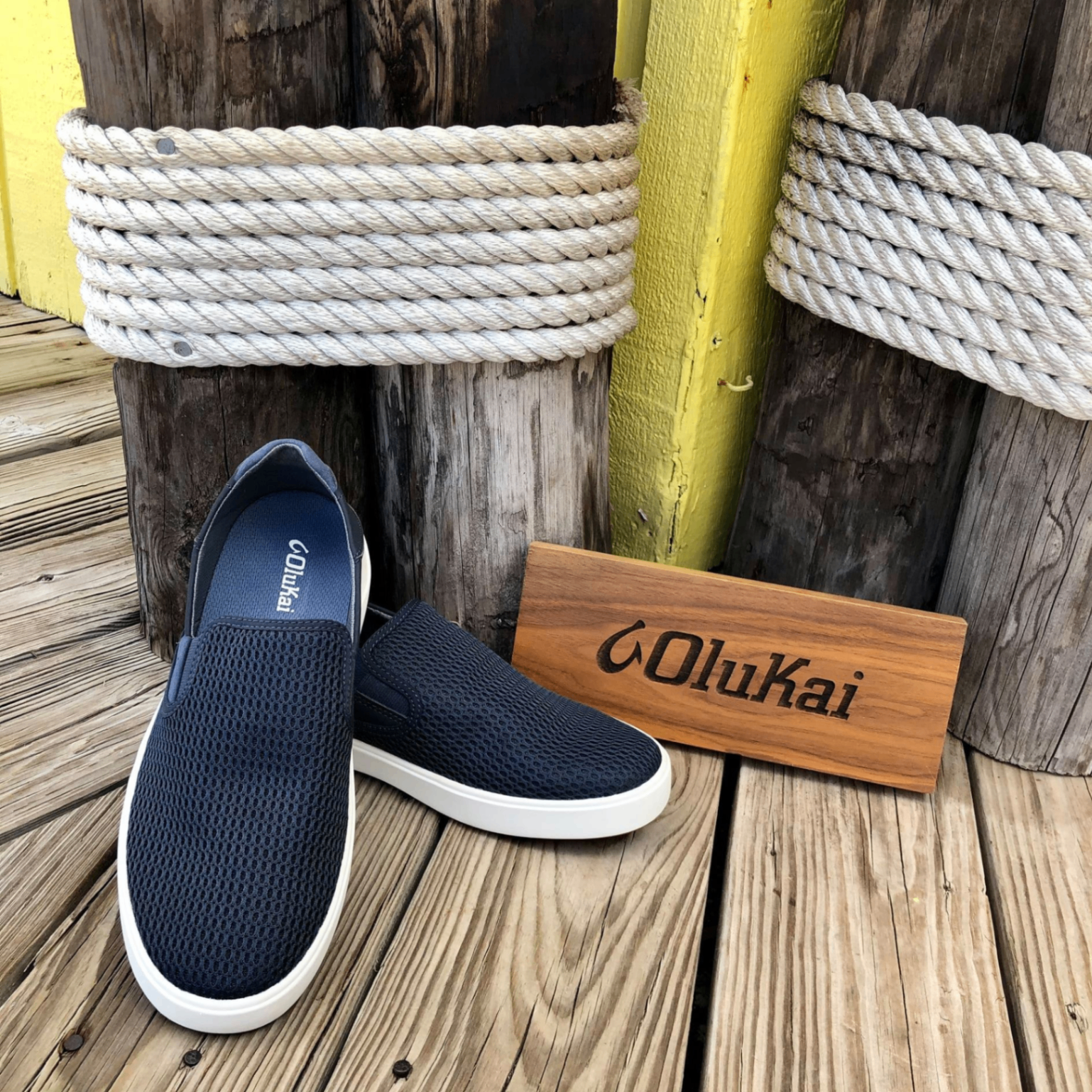 OluKai Pehuea navy mesh slip-on sneakers on a weathered dock with marine rope and yellow piling