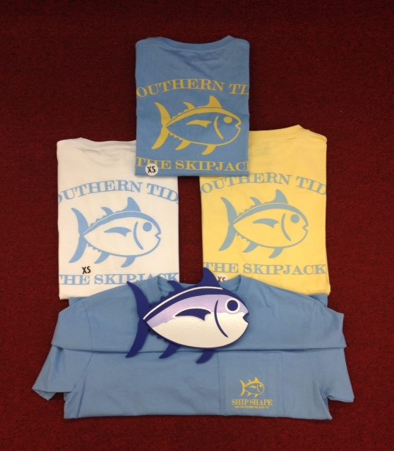 Folded Southern Tide Skipjack tees in blue, white and yellow arranged around a wooden fish logo